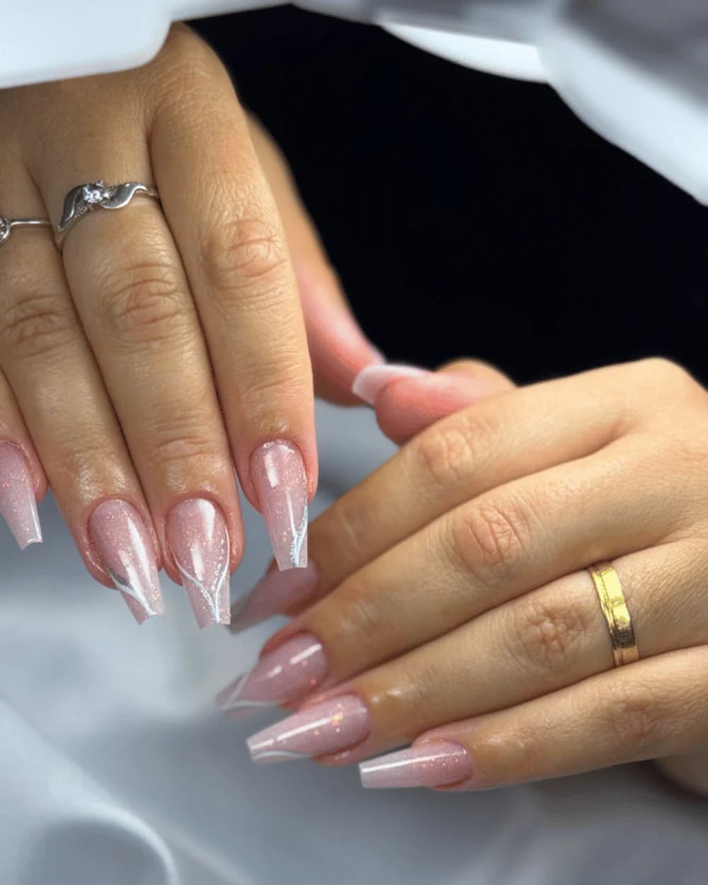 Unhas em formato bailarina com acabamento nude perolado e detalhe em branco, fotografadas sobre tecido claro.
