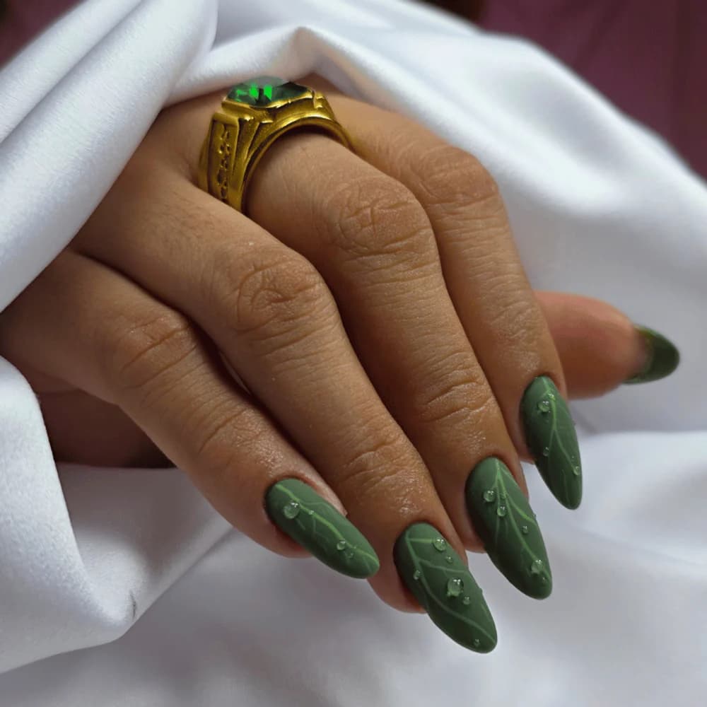 Unhas amendoadas em verde fosco com nervuras e gotas em relevo, ao lado de um anel dourado sobre tecido branco.